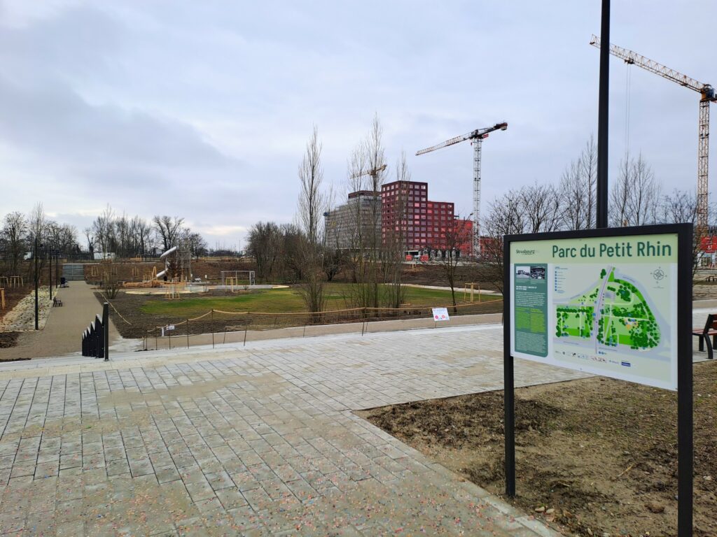 Vue du parc du petit rhin depuis l'arrêt Starcoop : on voit un parc en creux, dans lequel on descend par des escaliers. Le panneau du parc avec des arbres en contrebas en premier plan, de l'herbe en train de pousser dans un terrain humide (c'est l'hiver, le ciel et gris) et des bâtiments livrés et en construction dans le fond, du côté de Starlette