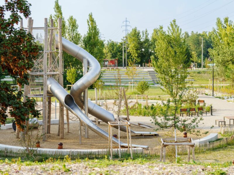 Des jeux pour enfants avec une tour et un toboggan sur un sol en copeaux de bois bordé par un banc en béton en arc de cercle. Au premier plan et au fond des arbres et chemins.
