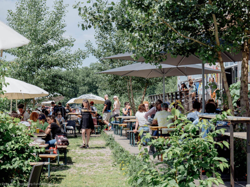 Festin du Turfu sur la terrasse du Phare Citadelle, des personnes assises sur des tables de brasserie sous des parasols au bord de l'eau, le paysage est arboré