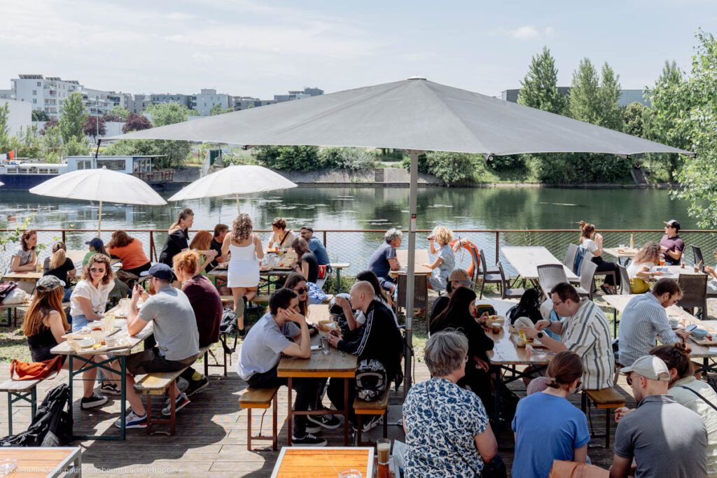 Festin du Turfu sur la terrasse du Phare Citadelle, des personnes assises sur des tables de brasserie sous des parasols au bord de l'eau, le paysage est arboré, on voit l'eau et le garde corps