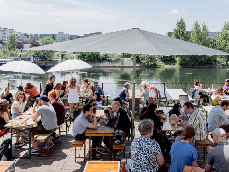 Festin du Turfu sur la terrasse du Phare Citadelle, des personnes assises sur des tables de brasserie sous des parasols au bord de l'eau, le paysage est arboré, on voit l'eau et le garde corps
