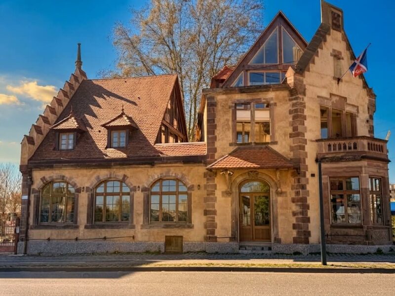 Une bâtisse restaurée avec un toit à double pan, des chiens assis, une terrasse ouverte à l'étage et des pignons à créneau. De grandes fenêtres longent la rue et un balcon surplombe une oriel.