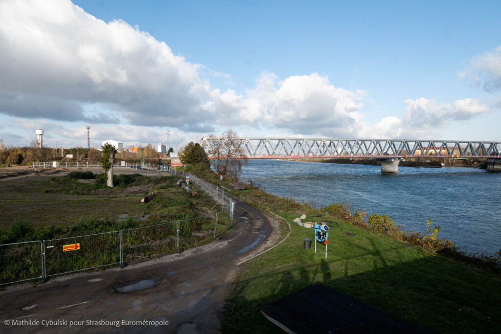 Accès au city-stade provisoire. Vue sur l'emprise de la future promenade du Rhin, une route en gravier longe les chantiers de le Cour des Douanes et le fleuve. 
