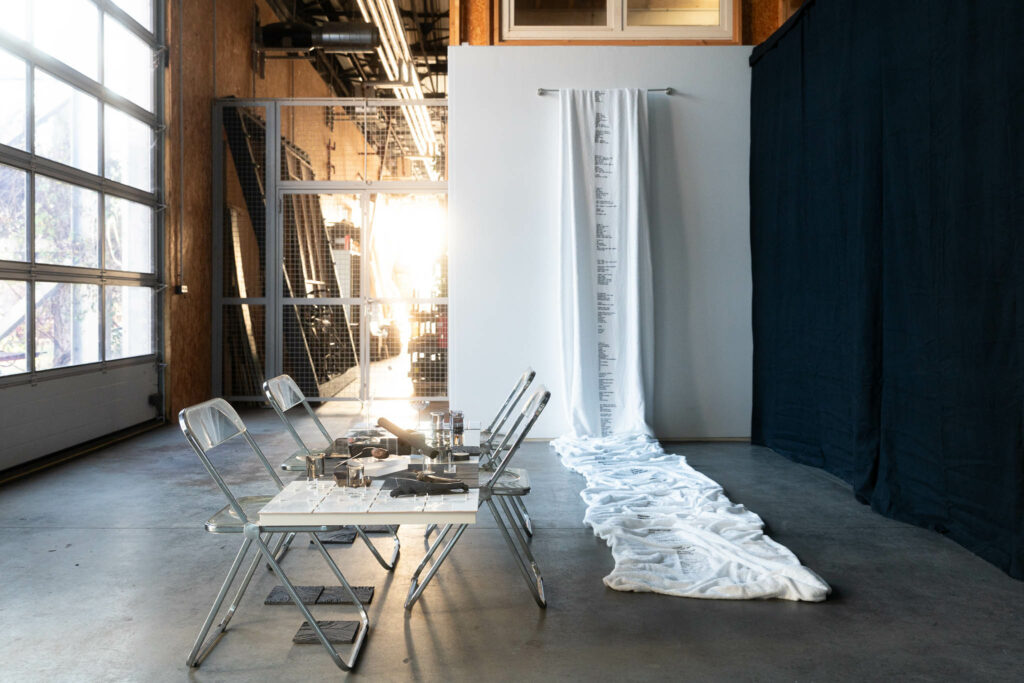 Un tissu imprimé d'un texte pend d'un mur et s’épand le long d'une table. Une installation de Sarai Rose Duke et Paula Beck dans le cadre de la Regionale 26 à Garage Coop