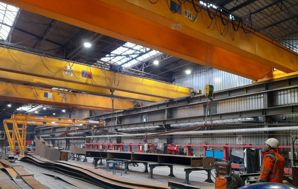 Vue sur les modules de traverse qui font 9m de long pour 50 à 60cm de large et marquent une ondulation. On voit un ouvrier en coin de photo qui donne l'échelle, ce qui permet d'estimer un entrepôt de 15 à 20m  de haut.