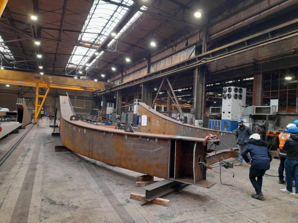 On voit un groupe de personne qui inspecte le début de 2 arches en acier de plus de 5m de long et qui vont de 1m à 3m de hauteur