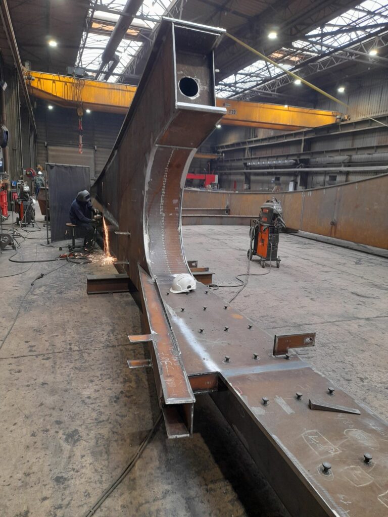 Un soudeur à l’œuvre sur un début d'arche métallique qui culmine à 3m de haut.