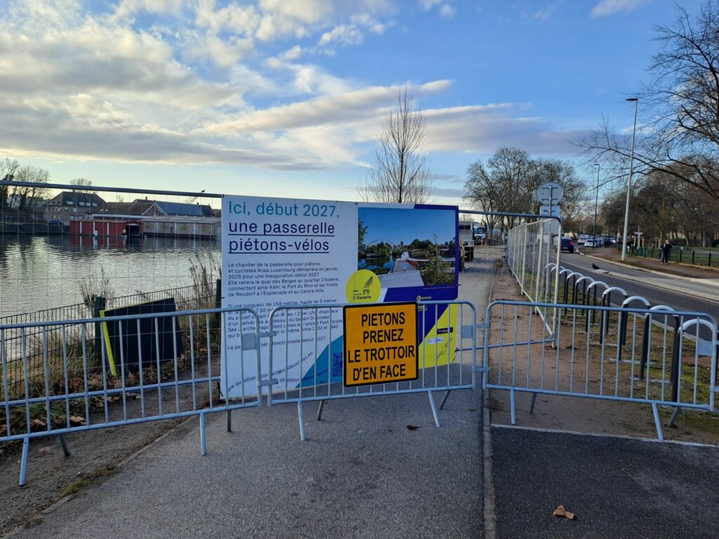Panneau piétons prenez le trottoir d'en face fixé sur des barrière qui clôturent le chantier. Derrière, un panneau annonce : Ici, début 2027, une passerelle piérons-vélos?