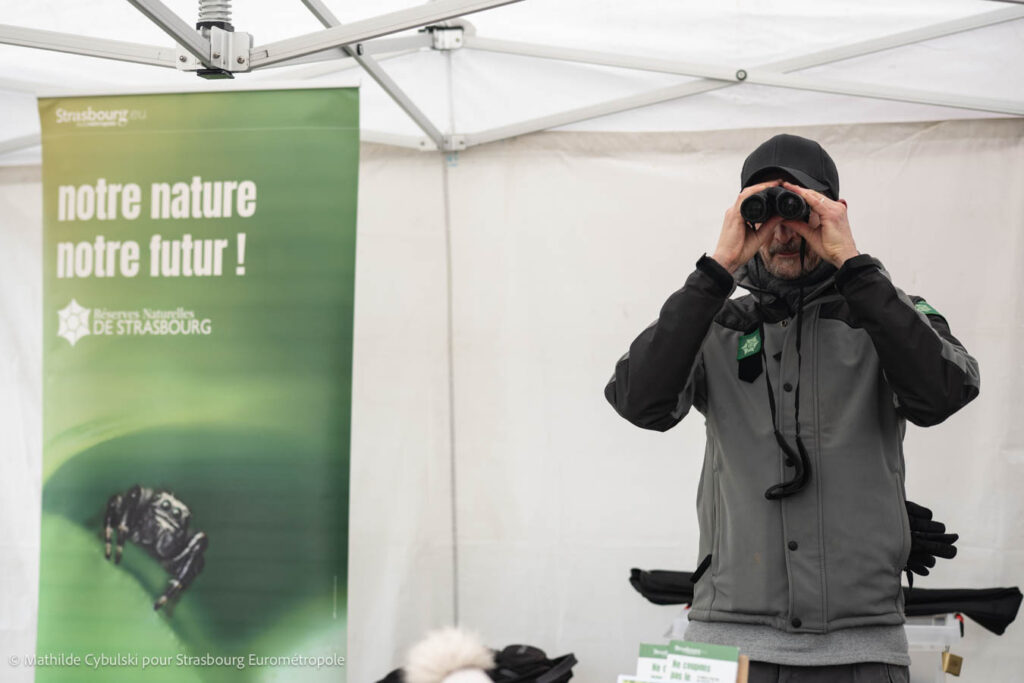Une personne pose avec des jumelles à côté d'un kakemono  "Notre Nature notre Futur" des réserves de la Ville de Strasbourg