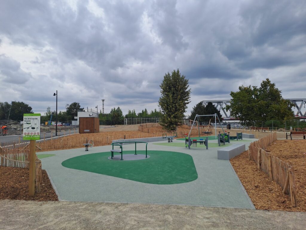 Une aire de jeu pour enfants avec un sol souple amortissant vert claire et vert foncé autour des jeux : un tourniquet, un jeu d'équilibre, une balançoire et une bascule (tape-cul). On voit des arbres et le pont de la voie de chemin de fer en fond