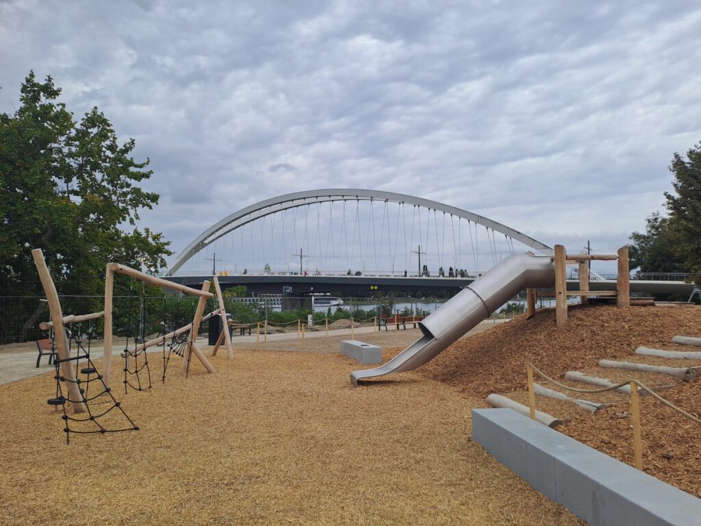 Une aire de jeux pour enfants avec un sol en copeaux de bois, une structure pour grimper et un toboggan installé sur une butte, on voit le pont du tram et le Rhin en fond