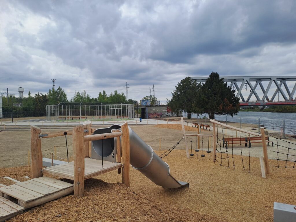 Une aire de jeux pour enfants avec un sol en copeaux de bois, une structure pour grimper et un toboggan installé sur une butte, on voit le city stade, le pont de la voie de chemin de fer et le Rhin en fond