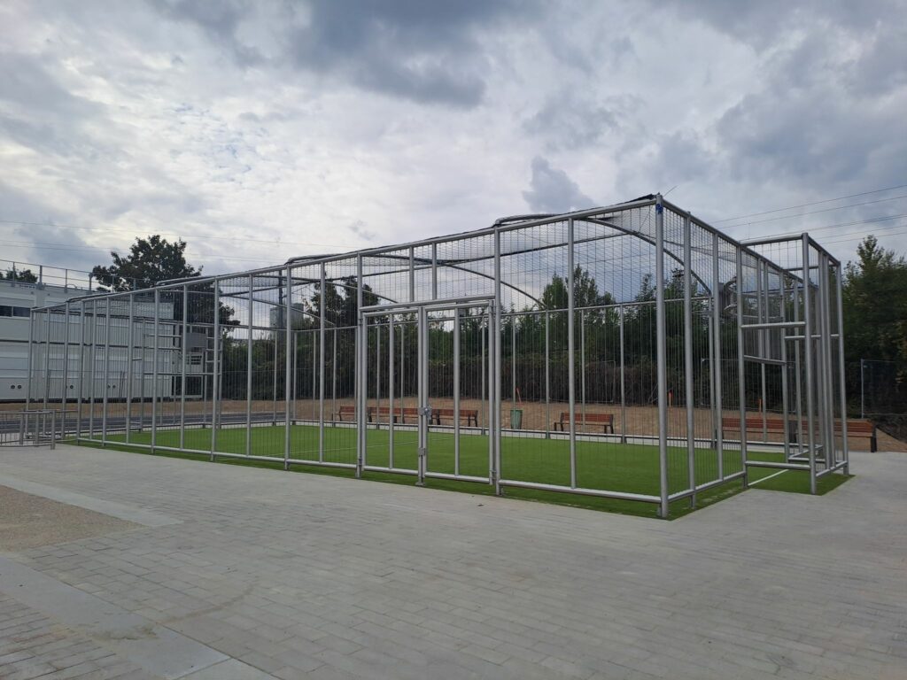 Un city stade fermé avec un filet pare ballons au plafond et des grilles pare ballons sur une structure en inox autour, avec un sol en synthétique vert et sable, un panneau de basket et les bâtiments modulaires (type algecco) de chantier en fond