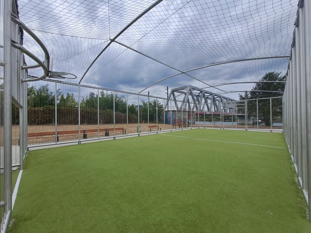 Un city stade fermé avec un filet pare ballons au plafond et des grilles pare ballons sur une structure en inox autour, avec un sol en synthétique vert et sable, un panneau de basket et le pont de la voie de chemin de fer en fond