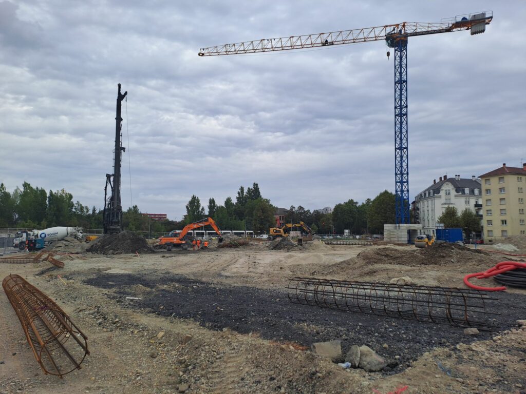 Un chantier avec des fers à béton en premier plan, une grue et une foreuse (machine à pieux) en second plan