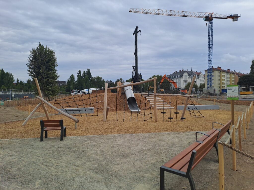 Une aire de jeux pour enfants avec un sol en copeaux de bois, une structure pour grimper et un toboggan installé sur une butte, on voit un chantier avec une foreuse, une grue et des engins en fond, du côté de la cité loucheur