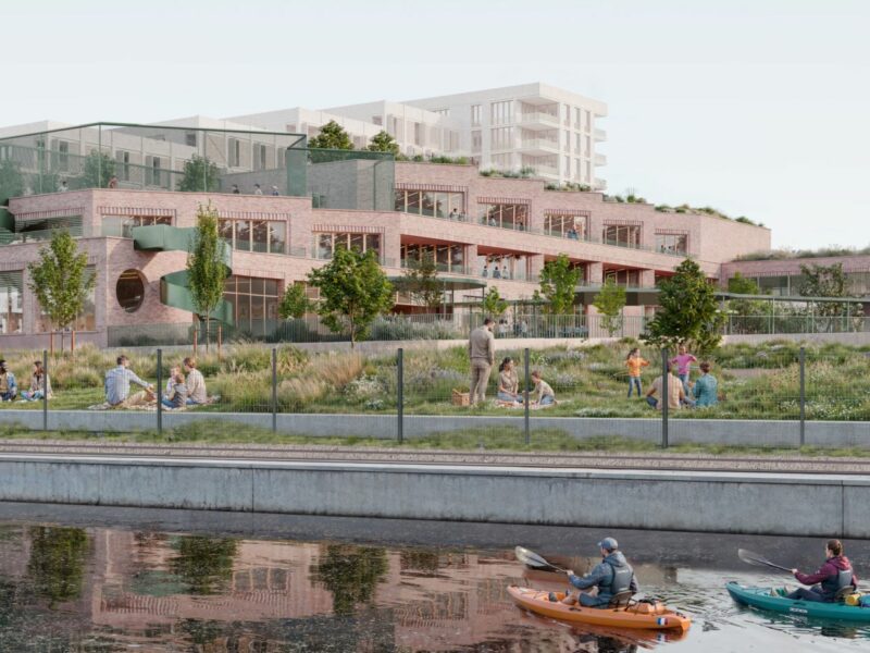 On voit un bâtiment de 2 étages en briques avec des façades ouvragées au bord de l'eau. Des personnes en kayak navigue sur le bassin au premier plan